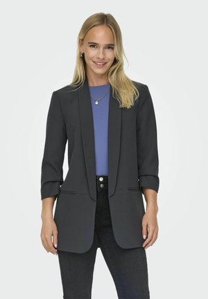 Jeune femme aux longs cheveux blonds portant un blazer noir, un haut bleu, un jean noir et un collier de perles, debout et souriante sur un fond blanc.