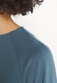 Haut en tissu bleu doux avec un col rond et des coutures subtiles aux épaules. Texture lisse avec un léger éclat. Accents ou motifs visibles limités.