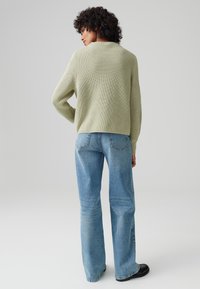 Hellgrüner gestrickter Pullover mit geripptem Muster, kombiniert mit hochgeschnittenen blauen Schlagjeans. Der Rücken des Models ist zu sehen, mit minimalem Hintergrund.