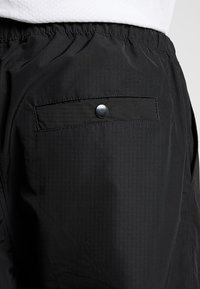 Black nylon shorts with an elastic waistband, featuring a back pocket secured by a silver snap button and a smooth, textured finish.