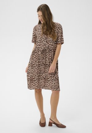 Naine, kes kannab põlveni ulatuvat lühikeste varrukatega leopardimustrilist kleiti, millel on nööriga talje ja pruunid madala kontsaga paelaga kingad, vaadates allapoole.