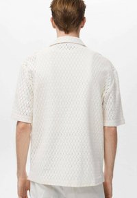 Camisa blanca de manga corta con patrón calado, cuello clásico y dobladillo recto. Textura ligera y diseño transpirable.