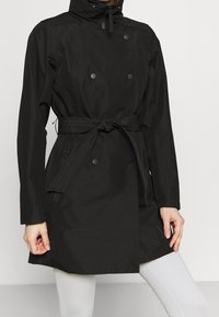Persona che indossa un trench coat nero con cintura, bottoni a pressione e tasche con zip, abbinato a pantaloni bianchi attillati.