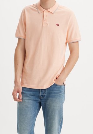 Mann trägt helles pfirsichfarbenes Kurzarm-Poloshirt mit kleinem rotem Levi's-Logo und hellblaue Jeans, eine Hand in der Tasche, vor schlichtem Hintergrund.