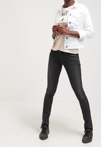 Chaqueta de denim blanca con cierres de botón, jeans negros ajustados y zapatillas negras, en capas sobre una camisa clara con texto gráfico.