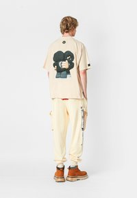 Camiseta oversized beige con un gran gráfico en la parte posterior, combinada con pantalones color crema y botas marrones. Diseño simple con un corte relajado.