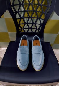 Par de mocasines de ante azul claro con plantillas color cuero colocados sobre una silla negra con respaldo geométrico contra una pared de azulejos amarilla y gris.