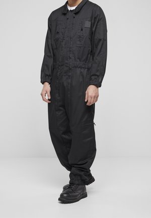 Homme portant une combinaison noire à manches longues avec des poignets élastiques et plusieurs poches zippées, associée à des bottes de combat noires, debout sur un sol gris.