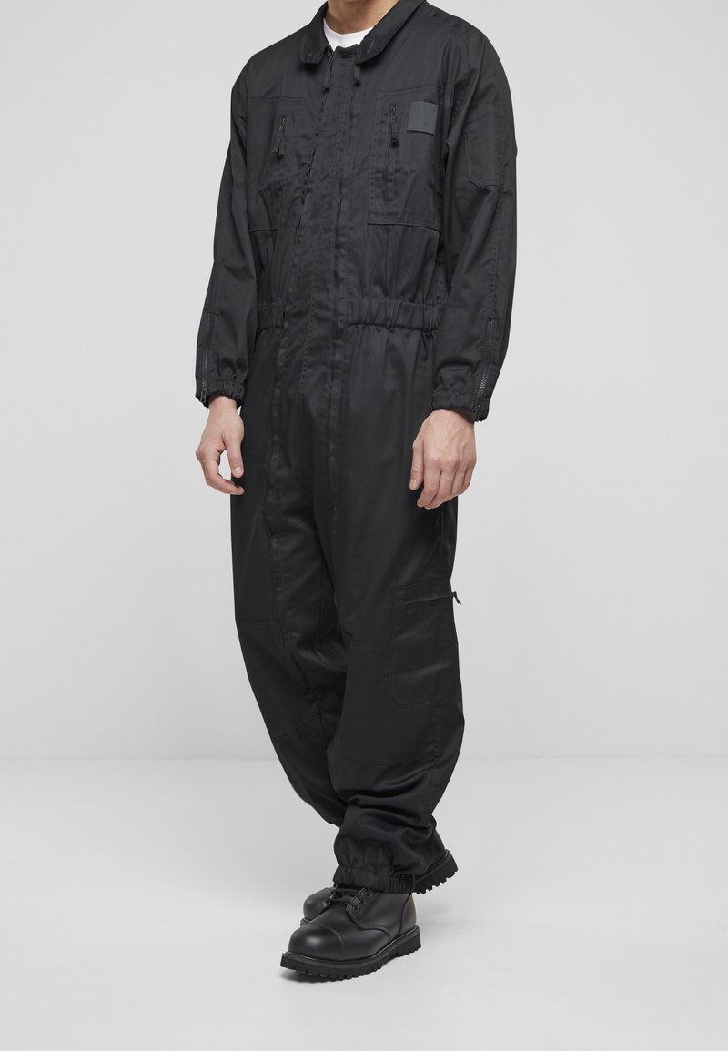 Homme portant une combinaison noire à manches longues avec des poignets élastiques et plusieurs poches zippées, associée à des bottes de combat noires, debout sur un sol gris.
