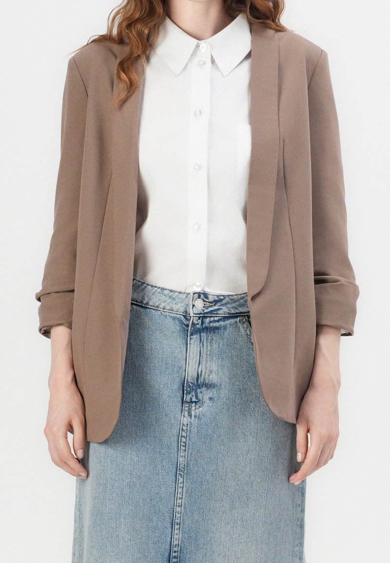 Mujer vestida con un blazer topo sobre una camisa blanca de botones metida en una falda de mezclilla azul claro, con las manos relajadas a los lados.