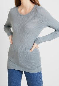 Hellgrauer Strickpullover mit einem strukturierten, atmungsaktiven Muster; hat einen weiten Ausschnitt und lange Ärmel; kombiniert mit blauen Jeans.