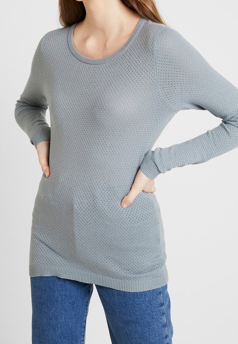 Hellgrauer Strickpullover mit einem strukturierten, atmungsaktiven Muster; hat einen weiten Ausschnitt und lange Ärmel; kombiniert mit blauen Jeans.