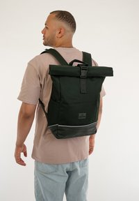 Fekete, felgörgő hátizsák tartós anyagból, elülső zsebbel, állítható pántokkal és fényvisszaverő díszítéssel az alján.