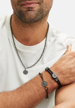 Schwarze Metal-Halskette mit Anhänger und passendem Handgelenksarmband; mit kunstvollen gravierten Designs. Über einem weißen T-Shirt getragen.