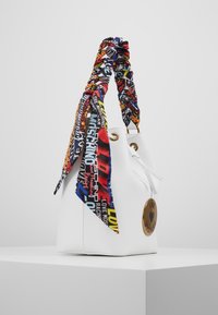 Vitt bärkorg med ett färgglatt mönstrat scarfhandtag, gyllene detaljer och en rund logotypaccent, med dragstringstängning.