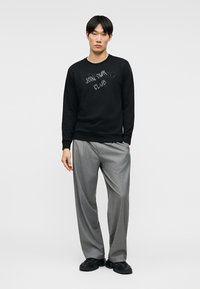 Homme portant un sweat-shirt noir avec le texte "JOIN OUR CLUB", un pantalon gris à jambes larges et des chaussures noires, debout devant un fond clair uni.
