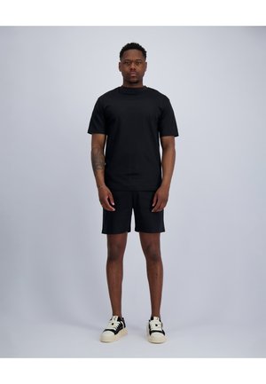 Man standing facing forward wearing a black t-shirt, black shorts, and black-and-white sneakers against a plain light background.