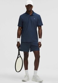 Granatowa koszulka polo i dopasowane szorty z lekkiego materiału, białe buty sportowe i opaski na nadgarstki; trzyma czarną rakietę tenisową.