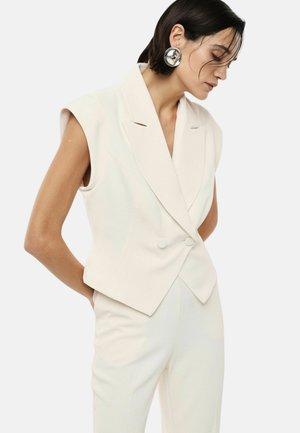 Donna che indossa un blazer bianco senza maniche con doppi bottoni, pantaloni abbinati e grandi orecchini rotondi d'argento, guarda verso il basso con le mani dietro la schiena.