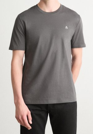 Homme portant un t-shirt uni gris foncé à manches courtes avec un petit logo blanc sur la poitrine et un pantalon noir, debout devant un fond clair.
