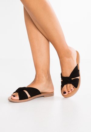 Fødder med sorte slip-on sandaler med åbne tæer, der har knyttede stofremme og naturligt farvede såler på en hvid gulvbaggrund.