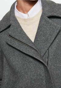 Abrigo de lana gris con solapas anchas y cremallera plateada expuesta, superpuesto sobre un suéter beige y una camisa blanca con cuello.