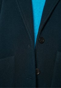 Tmavozelený vlnený kabát s klopovým golierom, dvomi prednými vreckami a jedným čiernym gombíkom na zapínanie. Pod ním je vidieť jasne modrý sveter.