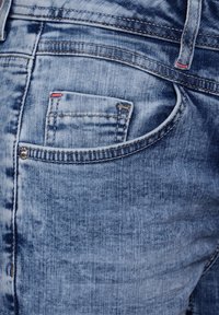 Los jeans de mezclilla de color azul claro cuentan con un bolsillo trasero que tiene una pequeña puntada de acento roja. La tela presenta una textura desgastada y detalles de costura típicos.
