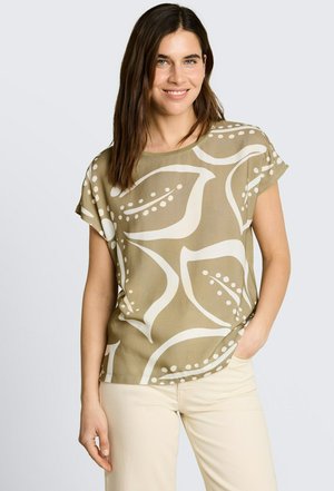 Uśmiechnięta kobieta z długimi brązowymi włosami, ubraną w beżowy top z dużymi białymi abstrakcyjnymi wzorami liści i kremowe spodnie, stojąca z jedną ręką w kieszeni.