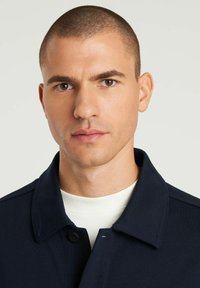 Marineblauer Mantel mit Kragen und Knopfverschluss, getragen über einem weißen Rundhals-Shirt. Glatter Stoff, minimalistisches Design mit feiner Textur.