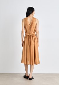 Robe midi en brun clair, avec un dos en V, des plis et une taille nouée. Fabriquée en tissu doux et fluide avec une coupe décontractée. Chaussures à talons noires portées.