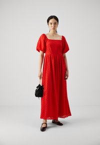 Rød maxikjole med pufferm, firkantet udringning og broderede detaljer. Båret med en sort taske og matchende mørke sko, sat mod en neutral baggrund.