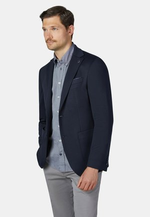 Mann trägt einen dunkelblauen Blazer über einem hellblauen Hemd mit Knöpfen und hellgrauen Hosen, steht mit Händen in den Taschen und schaut nach links.