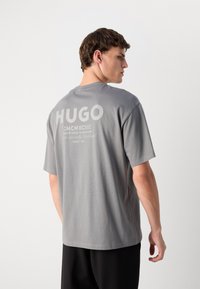 Grå bomull T-skjorte med korte ermer og rund hals. Har stor "HUGO" tekst i hvitt på ryggen, med mindre tekst under.