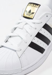 Vit lädersneaker med svarta tre ränder, guld-logotyp på tungan, texturerat gummitåhölje och vita snören. Solid design och klassisk form.