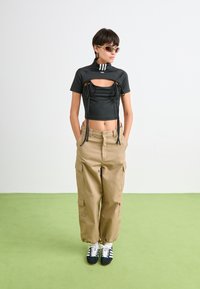 Henkilö, joka wear black cropped top with cutout, beige cargo-housut, aurinkolasit ja lenkkarit, seisoo vaaleanvihreällä lattialla valkoista seinää vasten.