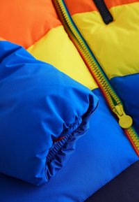Giacca imbottita caratterizzata da colori vivaci: arancione, giallo, blu. Texture in nylon morbido con polsini elasticizzati e un dettaglio della zipper colorato.