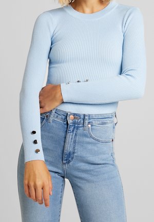 Kobieta ubrana w jasnoniebieski prążkowany top z długim rękawem, z srebrnymi guzikami na mankietach, oraz w jasnoniebieskie, wysokiej talii dżinsy.