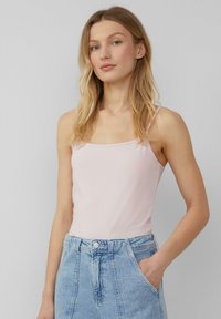 Frau mit langen blonden Haaren, die ein hellrosa ärmelloses Top und hochtaillierte blaue Denim-Jeans trägt, mit einer Hand in der Tasche.