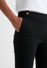 Czarne spodni z dopasowaniem o gładkiej fakturze, z złotymi guzikami i kieszeniami bocznymi. Pas jest usztywniony i płaski.