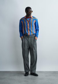 Mężczyzna w wzorzystej niebiesko-czerwonej koszuli na guziki, szerokich spodniach w drobne prążki, czarnych butach i okularach przeciwsłonecznych, stojący z rękami w kieszeniach.