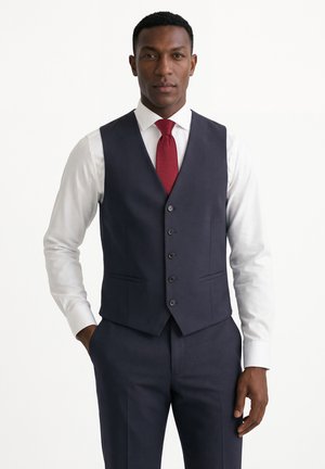 Mann trägt ein weißes Hemd, eine dunkelblaue Weste und Hose sowie eine rote Krawatte und steht mit einer Hand in der Tasche.