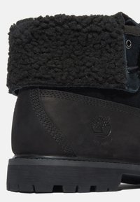 Botas de ante negro con un cuello de forro de lana negra texturizada, suela de goma y logo grabado. Presenta costuras reforzadas en los laterales.