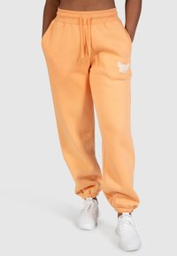 Orange sweatpants lavet af en bomuldsblanding, med elastisk talje, sidelommer og tætsiddende manchetter, der har et hvidt broderet design på det ene ben. Båret med hvide sneakers.