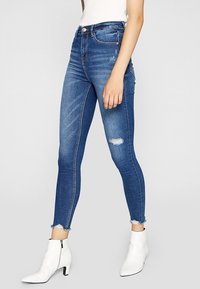 Högmidjade blå jeans med smal passform, med en slitning vid knäet, fransad fåll och subtila slitningar. Kombinerade med vita ankelboots.