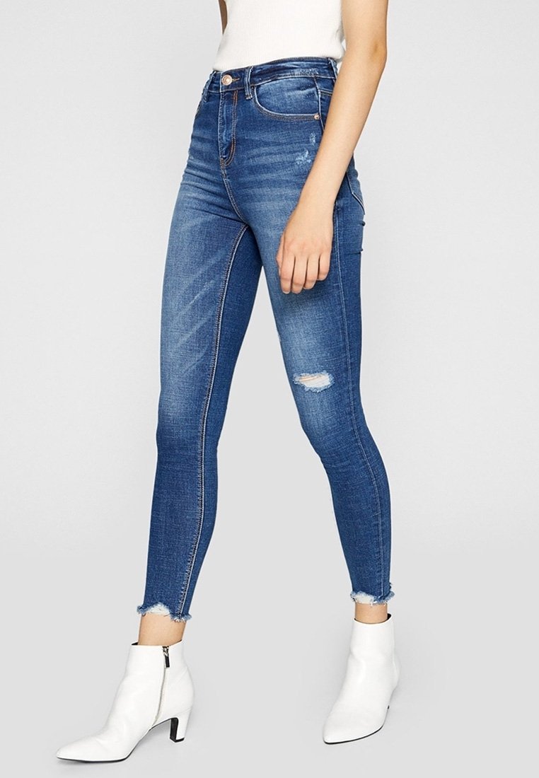 Högmidjade blå jeans med smal passform, med en slitning vid knäet, fransad fåll och subtila slitningar. Kombinerade med vita ankelboots.