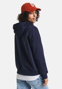 Navy blue hoodie with a relaxed fit and drawstring hood, paired with a white striped shirt, denim pants, and a red corduroy cap.
