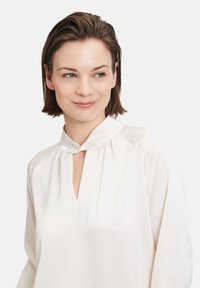 Blusa bianca con colletto alla mandarina e apertura a chiave sul davanti. Realizzata in tessuto liscio con una sottile texture e maniche lunghe.