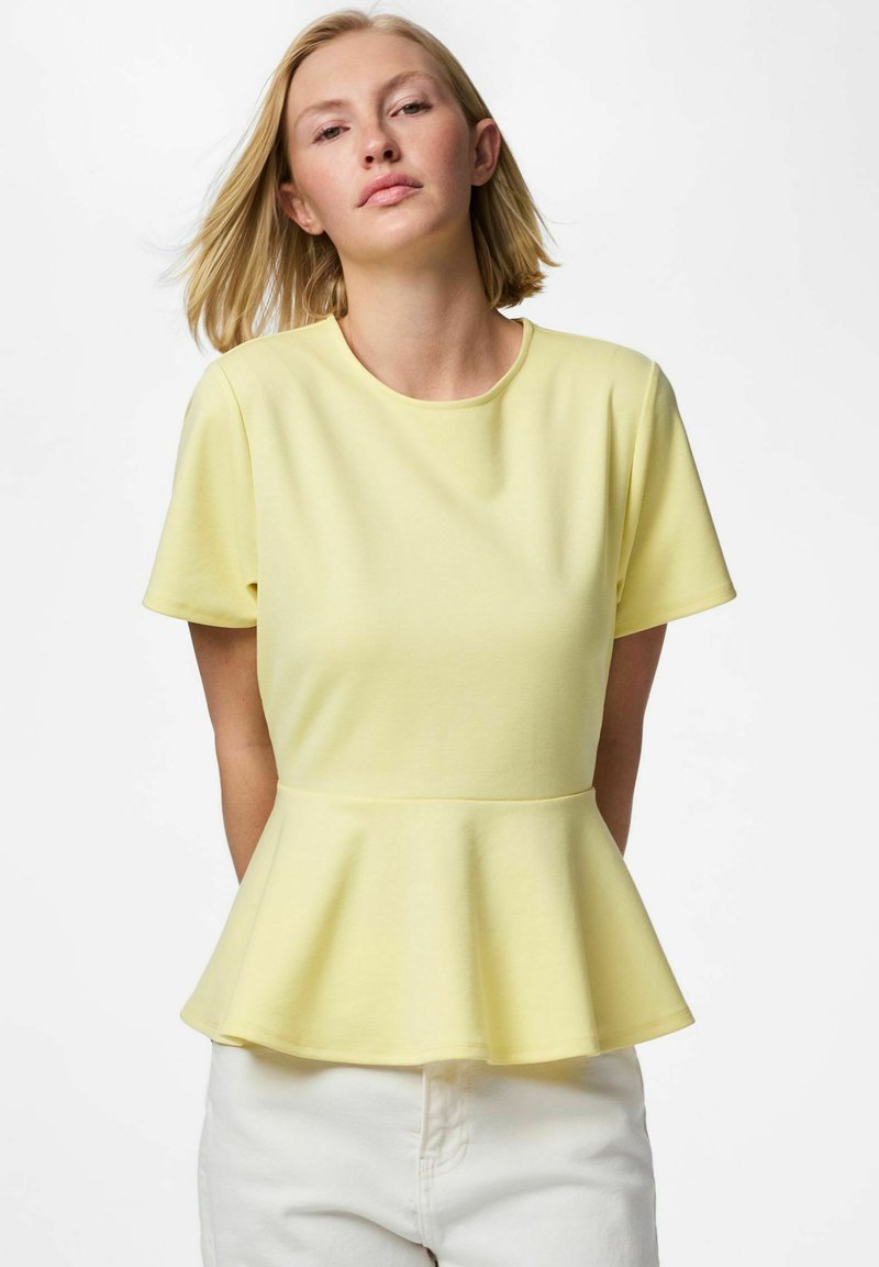 Femme aux cheveux blonds courts portant un haut peplum à manches courtes jaune pâle et un pantalon blanc sur un fond clair uni.