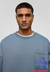 Blauwe longsleeve sweatshirt met een zak en meerkleurige branding. Zachte textuur, ronde hals, relaxte pasvorm en minimale details.